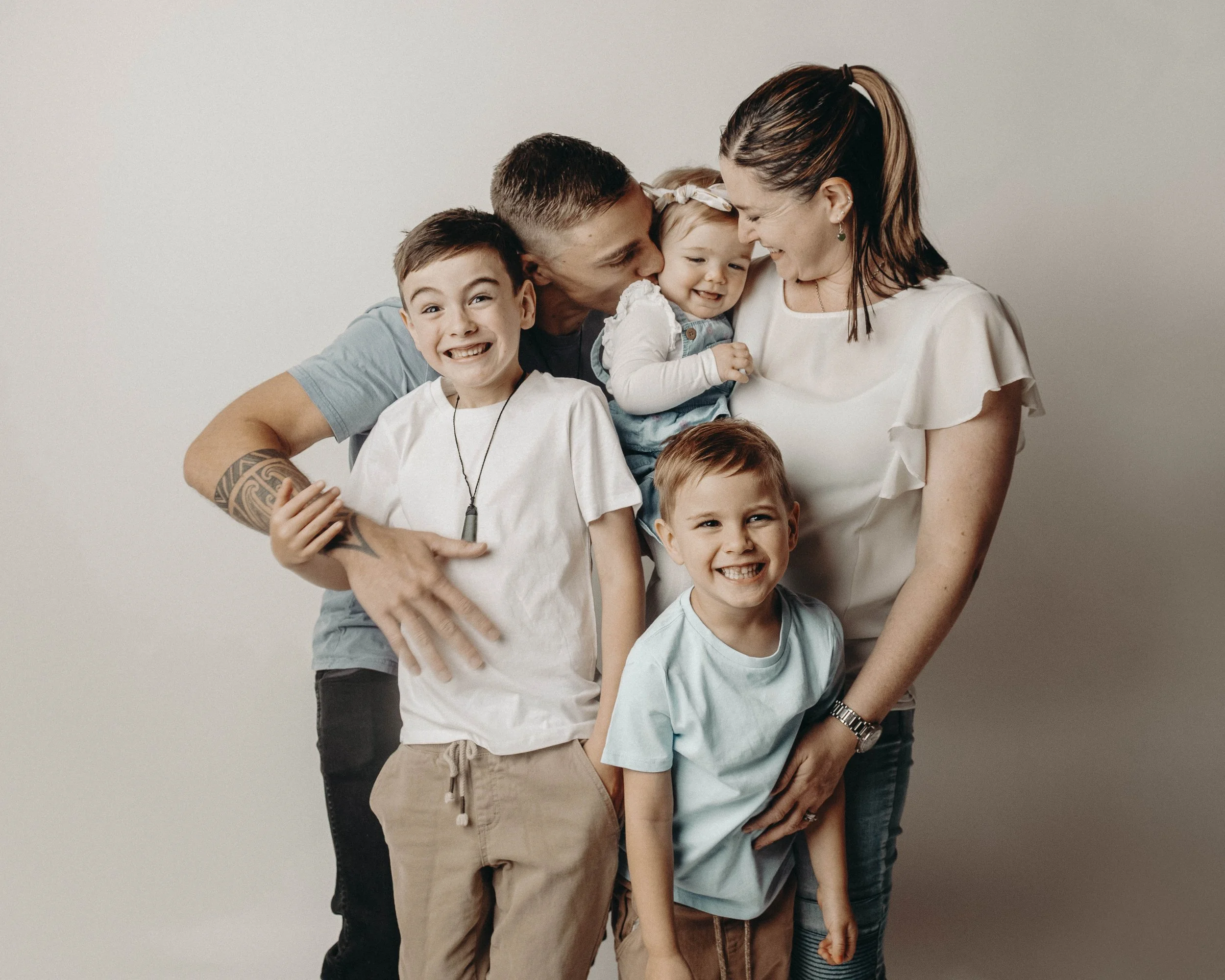 A woman with a ponytail and earrings is embracing and playing with four children, all smiling and hugging each other against a plain background.