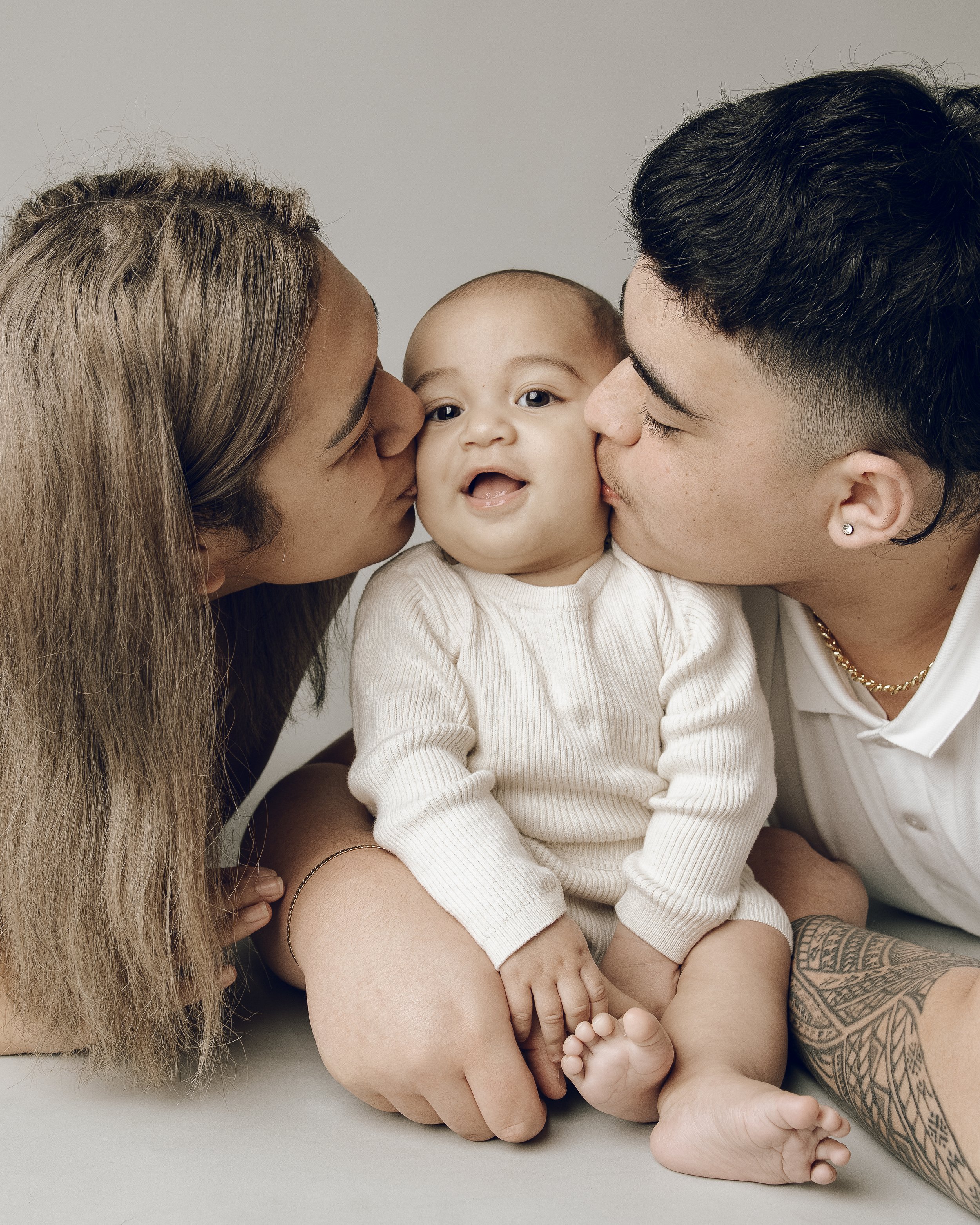 Close-up of a family moment with a baby being kissed on both cheeks by parents, one with long blonde hair and the other with short dark hair, showcasing love and affection.