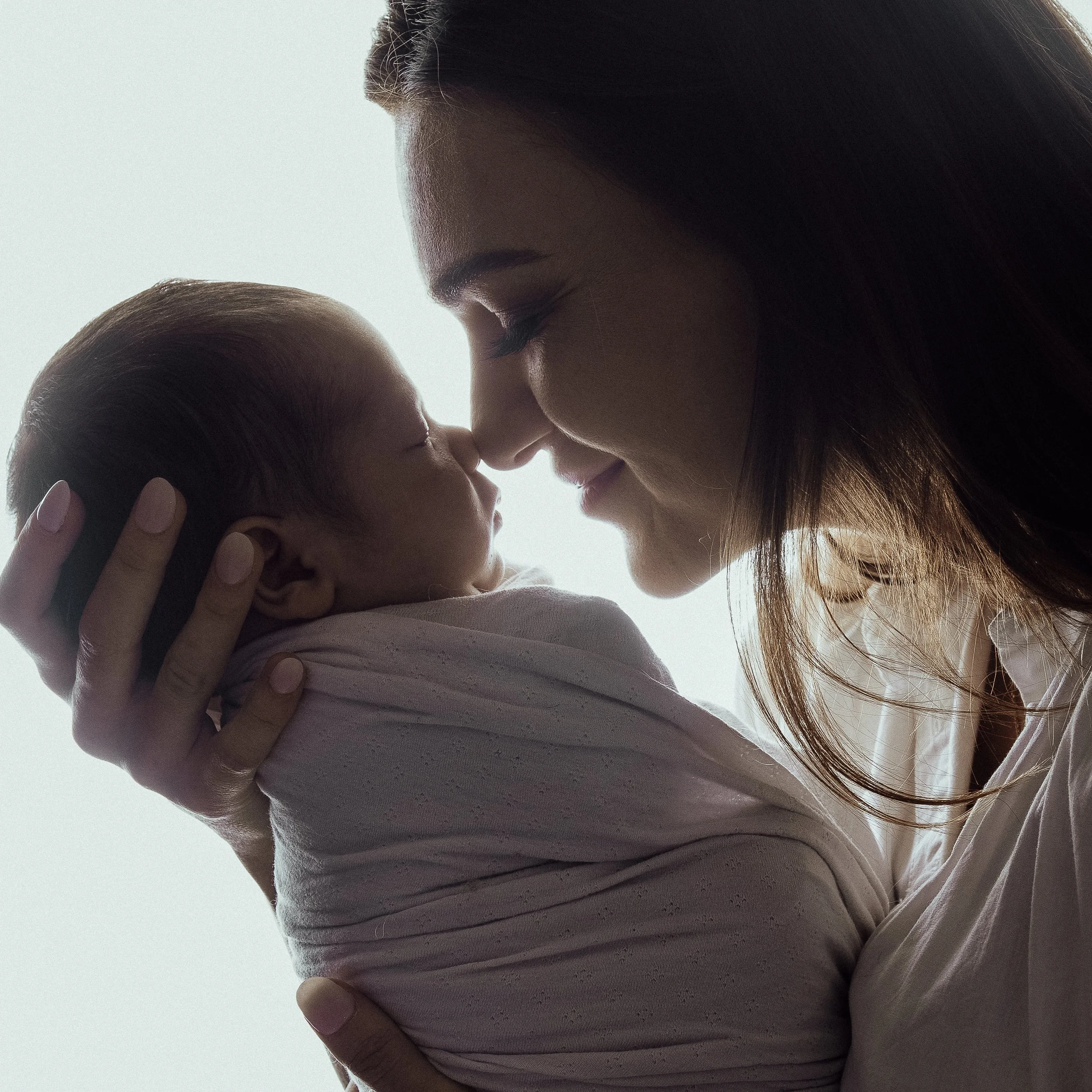 A close-up of a woman with long dark hair gently touching the forehead of a baby with short hair. They appear to be sharing a tender moment.