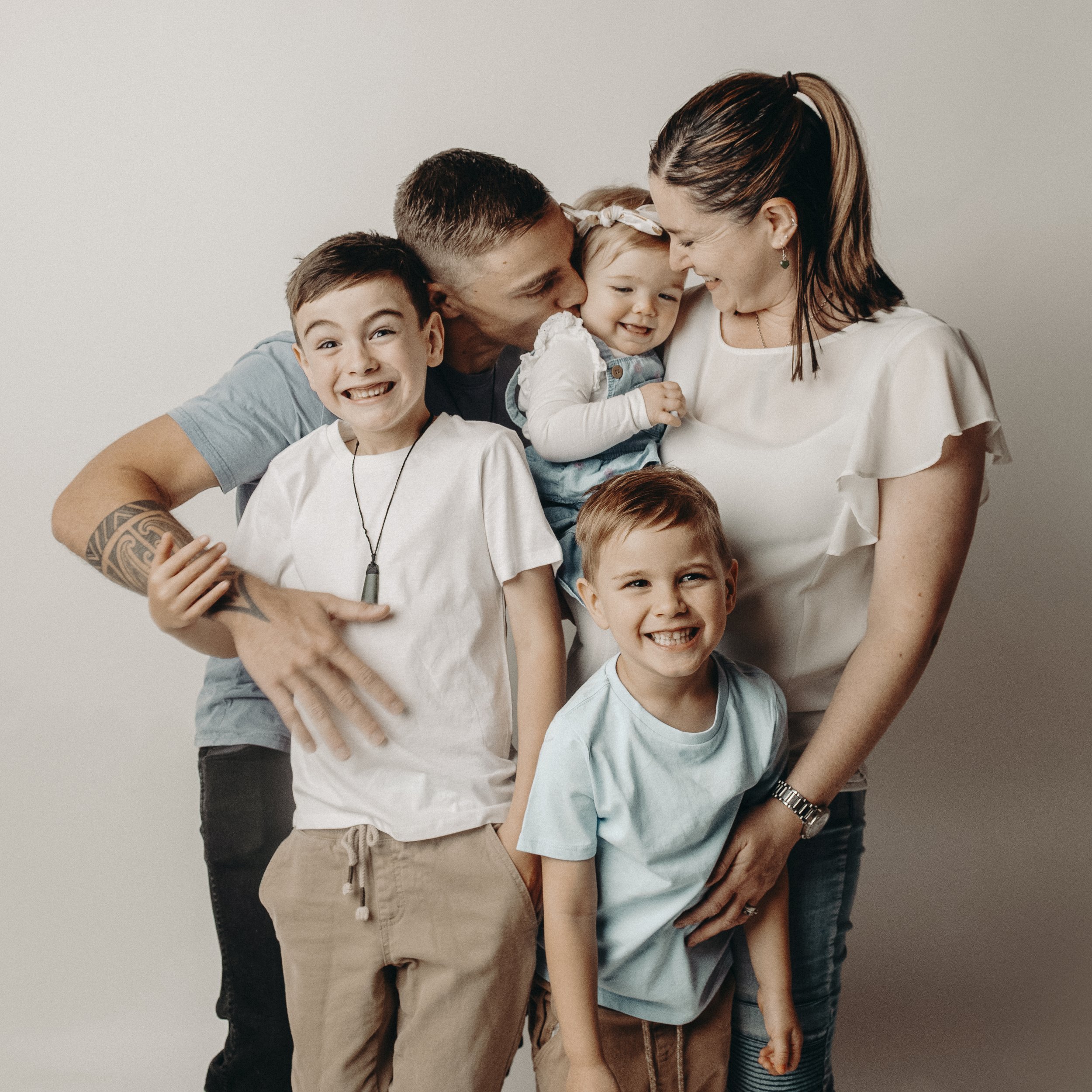 A group of six people, including two children, a man, and a woman, are smiling and hugging each other against a plain white wall. The woman has dark brown hair with light brown highlights, and the children have light brown hair. The group appears happy and affectionate.