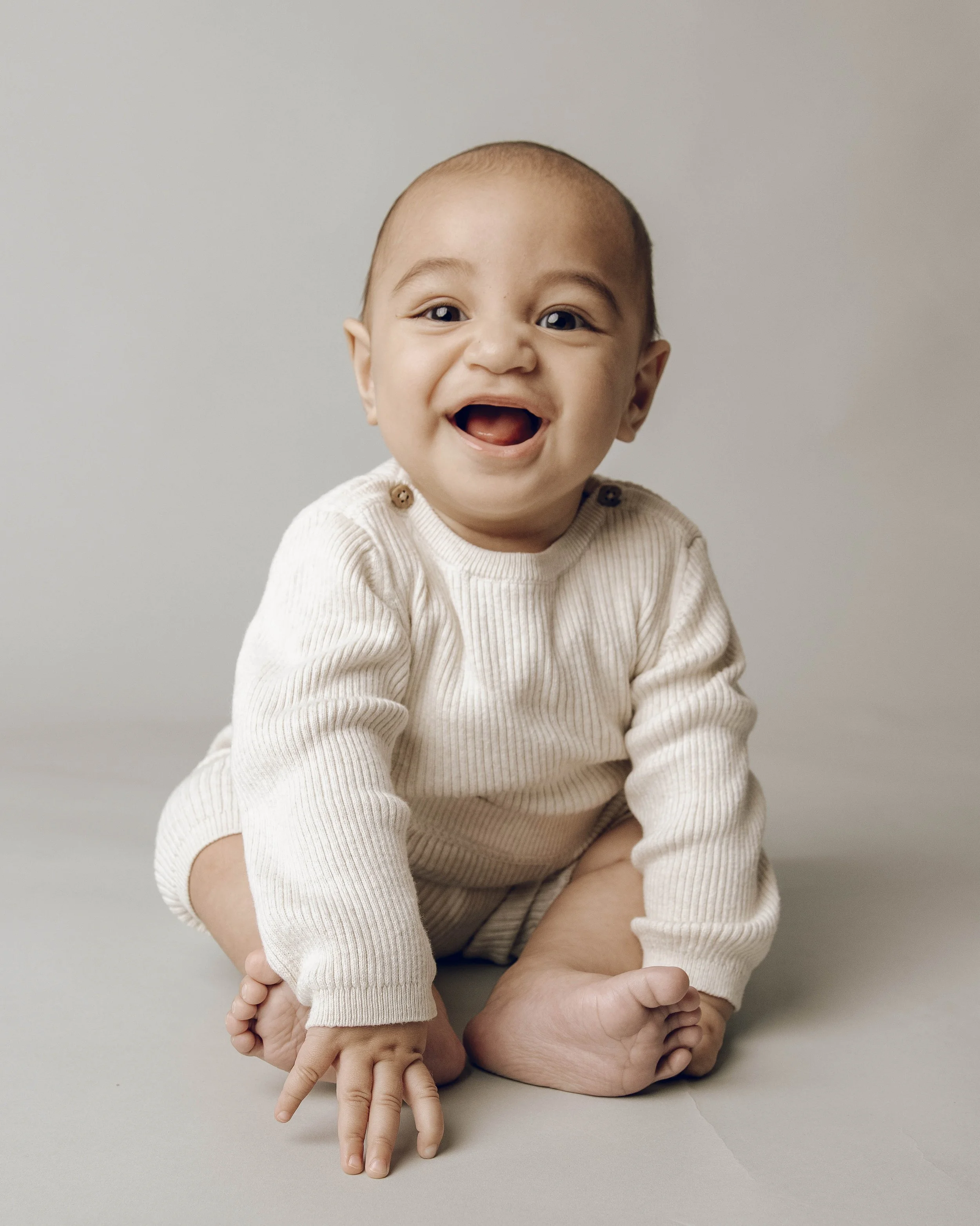 A smiling baby with light skin and dark eyes, sitting on a surface with a plain light background. The baby is wearing a cream-colored sweater with small buttons on the shoulder and is in a playful pose with one finger pointing down and the other hand