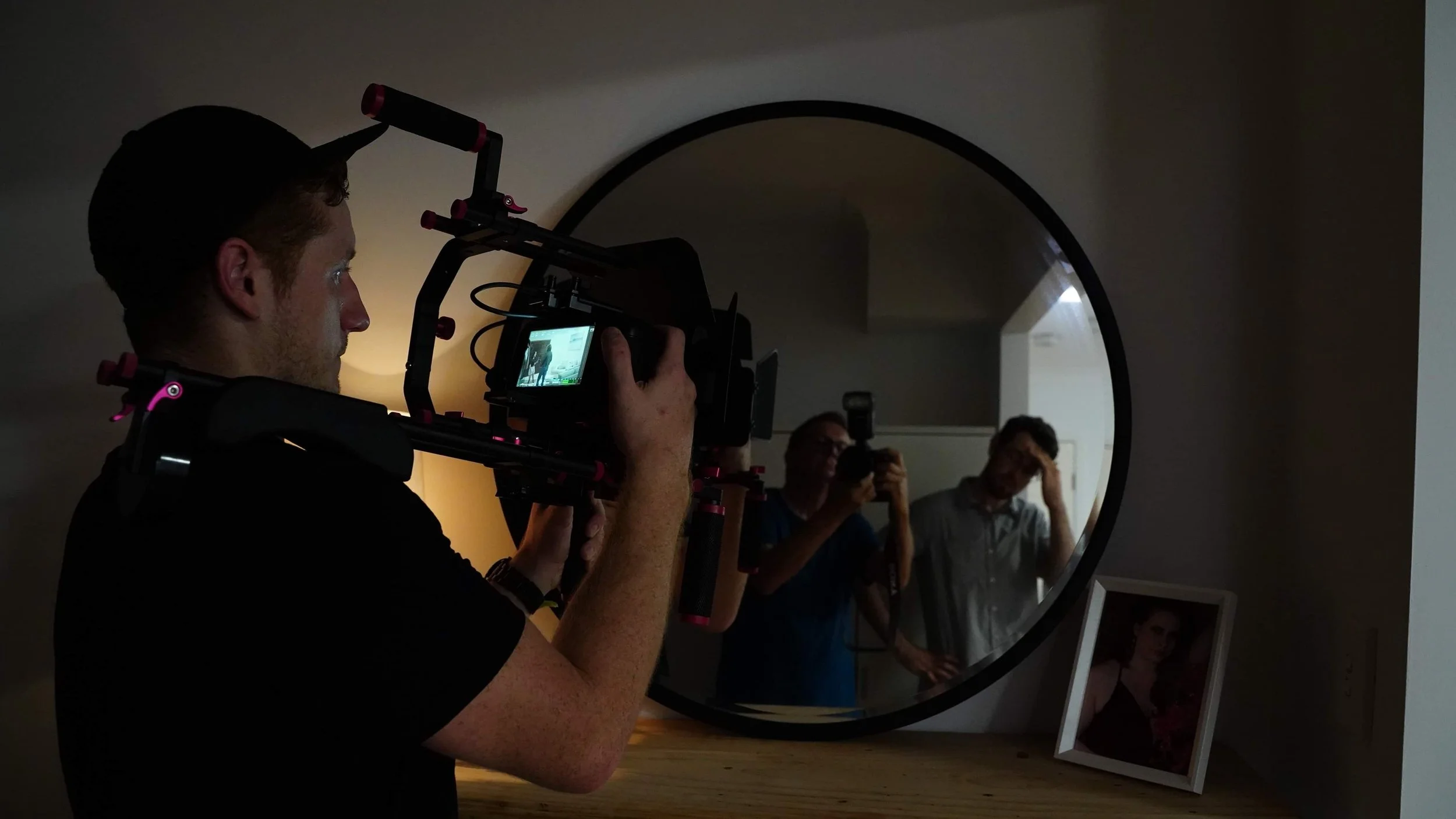 Person filming in front of a round mirror, with two people visible in the reflection and a framed photograph on the floor.
