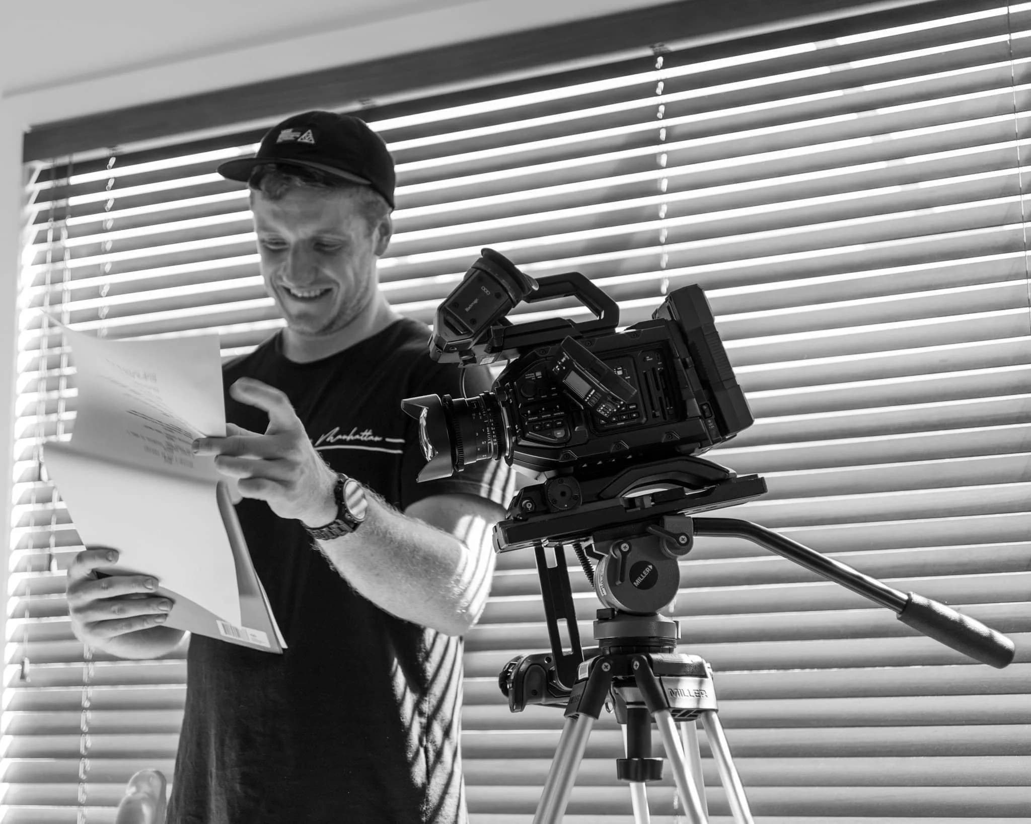 A man wearing a cap and t-shirt smiling while looking at a piece of paper, with a professional camera and tripod in front of him, in front of window blinds.