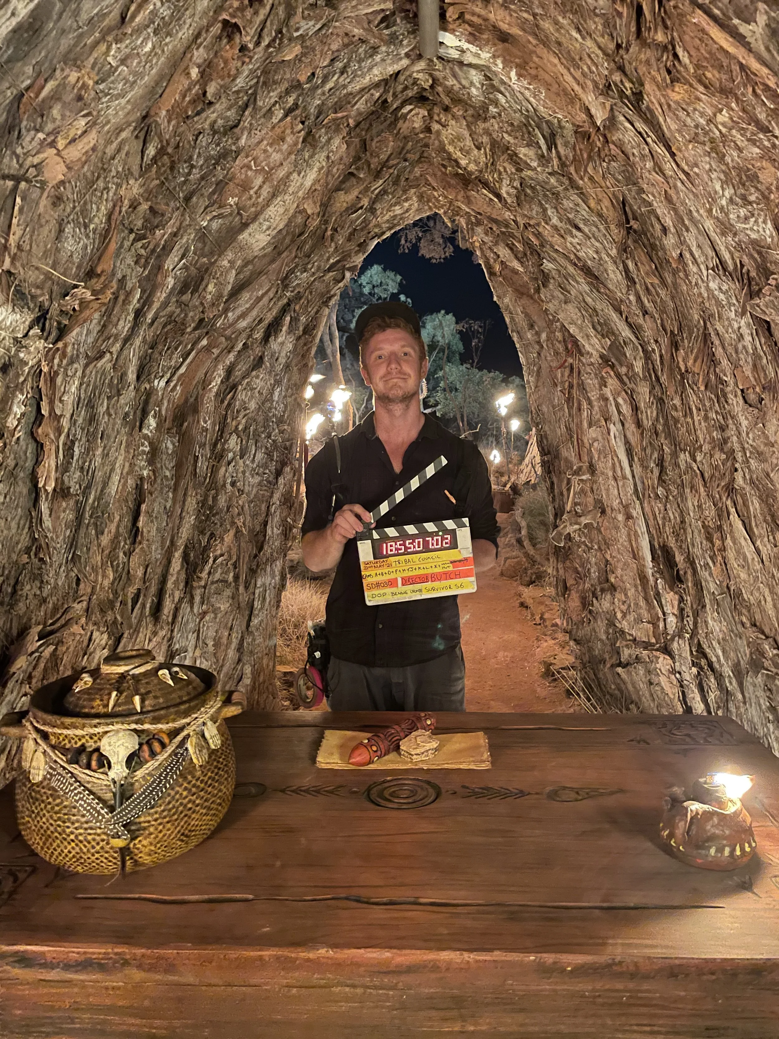 A man holding a clapperboard stands inside a large, natural tree tunnel at night. In front of him on a wooden table are traditional indigenous items, including a basket filled with jewelry, a carved wooden object, and a painted artifact. The background features trees and outdoor lights.