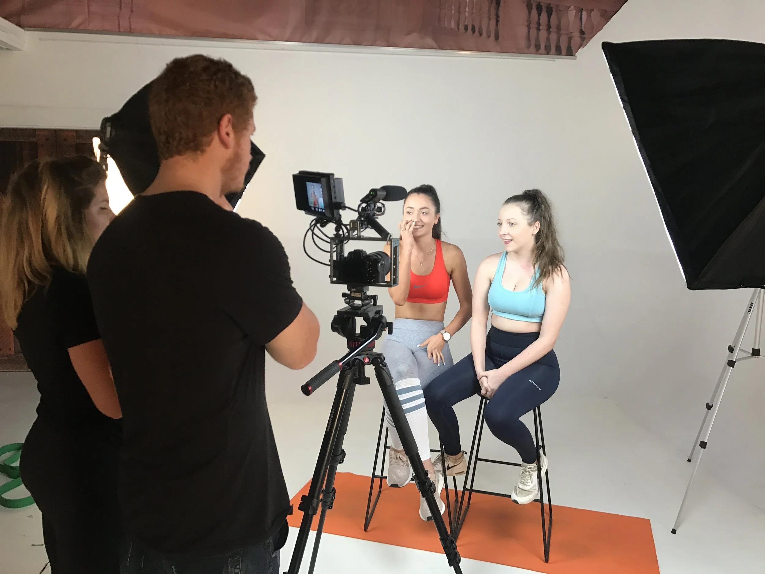 Two women in athletic wear sitting on stools in a photo studio, with lighting equipment around them, and a man operating a camera.