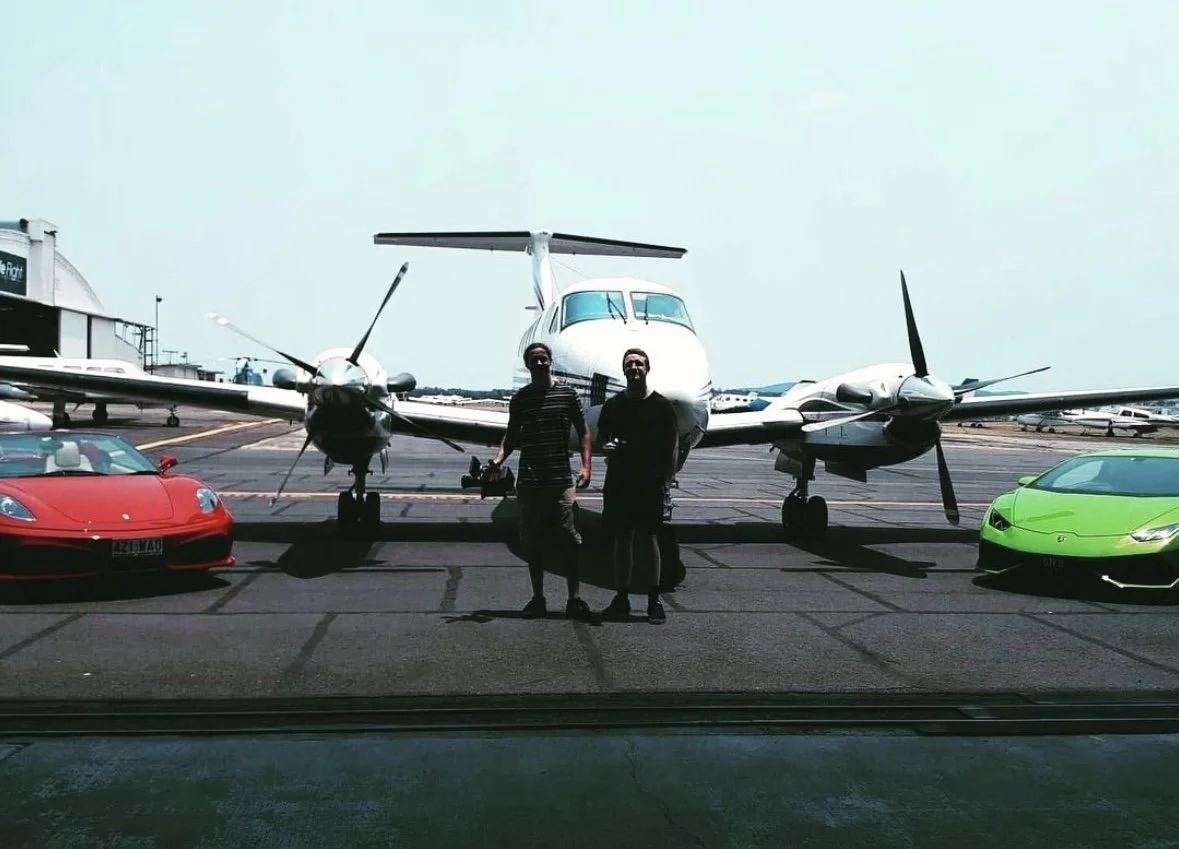 Two men stand on an airport tarmac in front of a small propeller airplane, flanked by a red sports car on the left and a green sports car on the right.