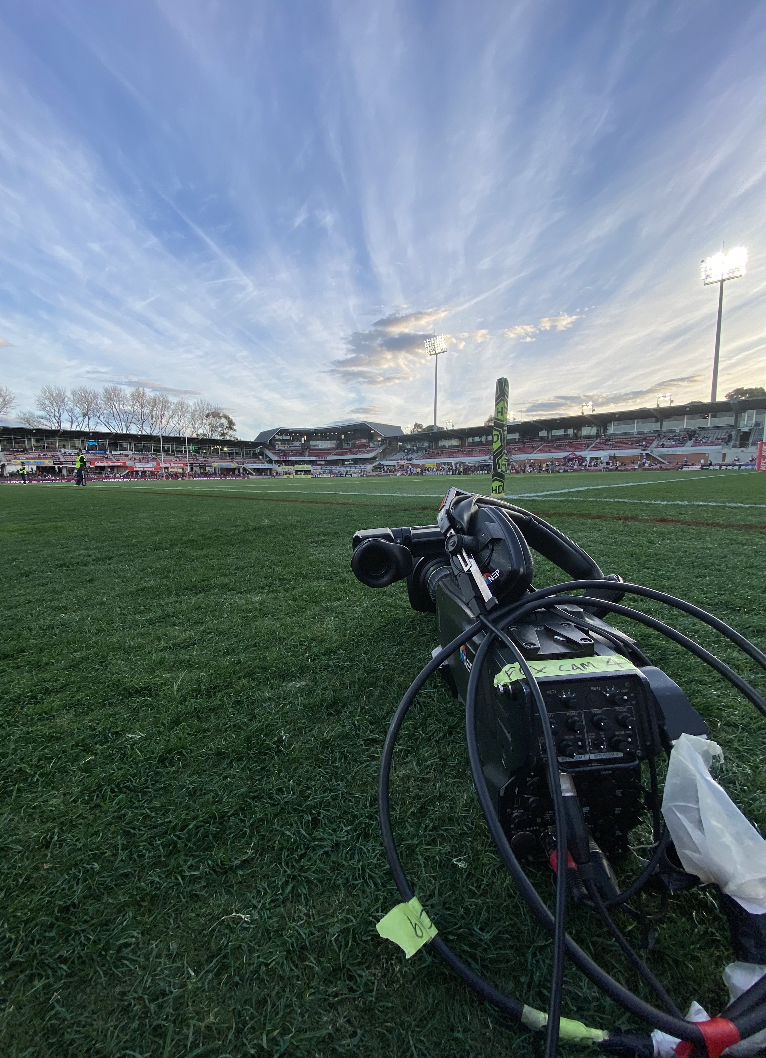View of a sports stadium field with floodlights, empty stands and a camera on the grass in the foreground, under a partly cloudy sky.