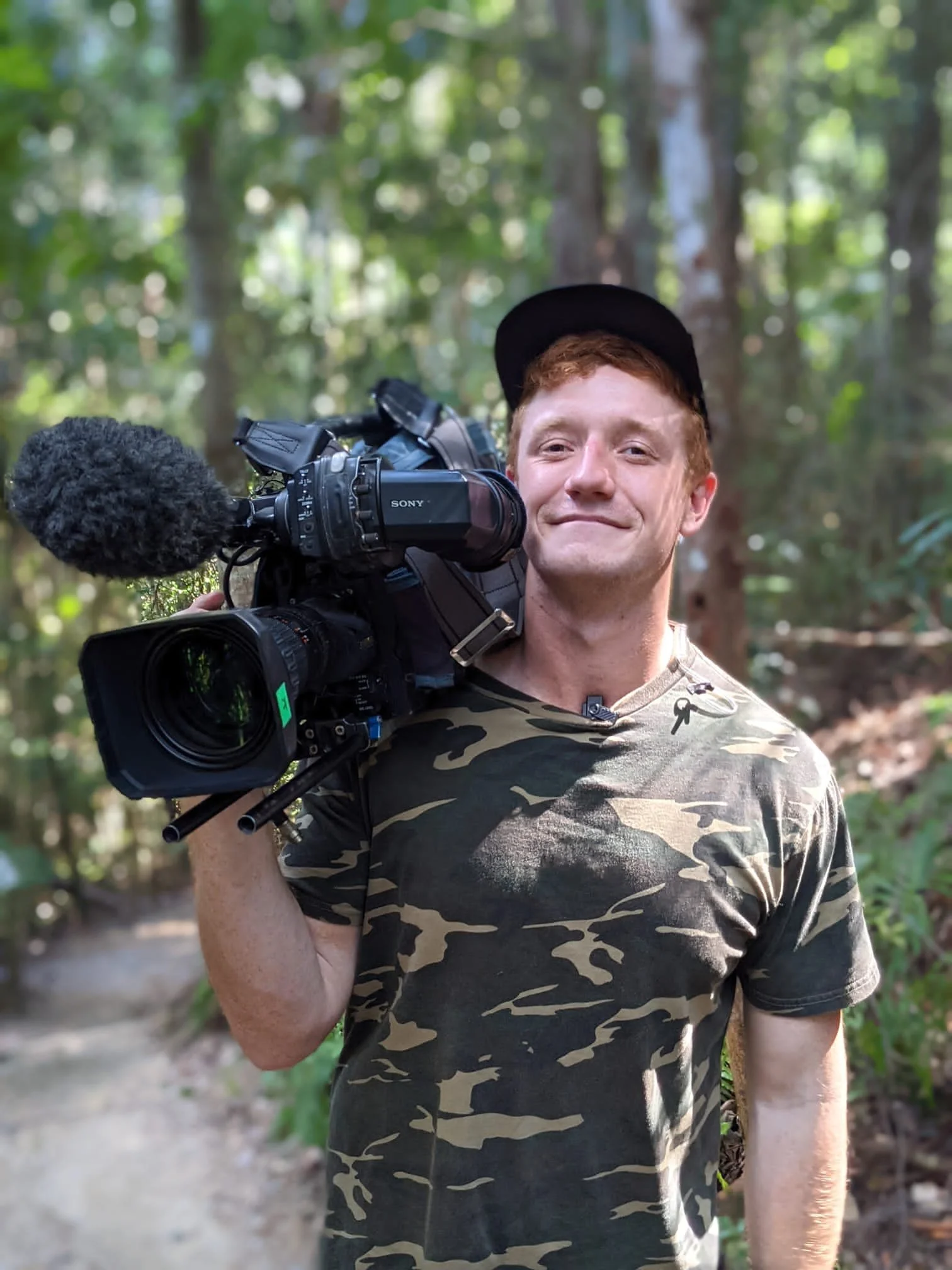 A young man with reddish hair and wearing a black cap, camouflage shirt, is smiling and carrying a professional video camera on his shoulder in a forest setting with trees and greenery.