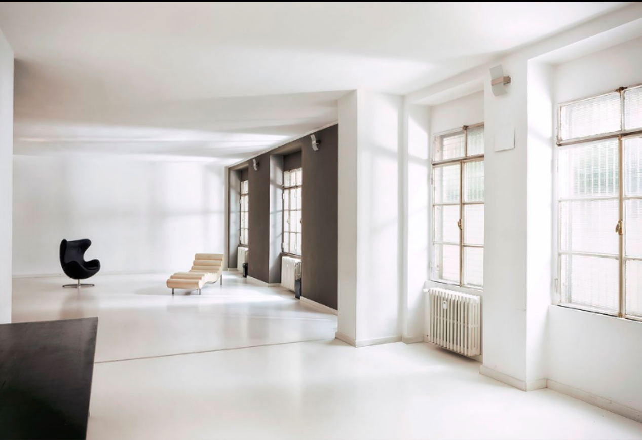 Interno di un loft moderno e minimalista con pareti bianche, grandi finestre con infissi di legno e radiatori, arredato con una sedia nera a forma di uovo, una chaise longue beige e un mobile scuro.