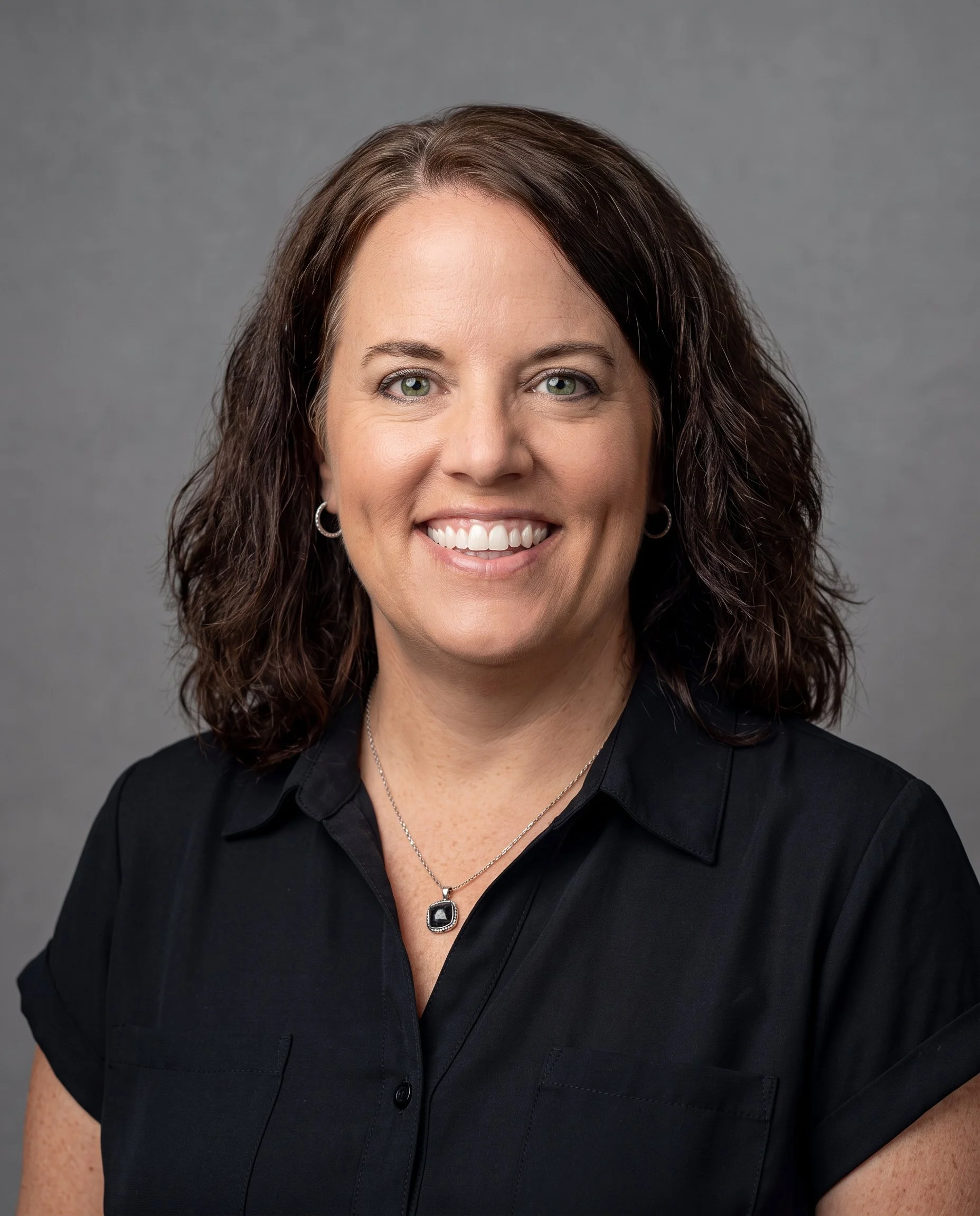 Close-up portrait of a smiling woman with brown, curly hair, wearing a black shirt, silver earrings, and a silver necklace with a black gemstone pendant, against a gray background.