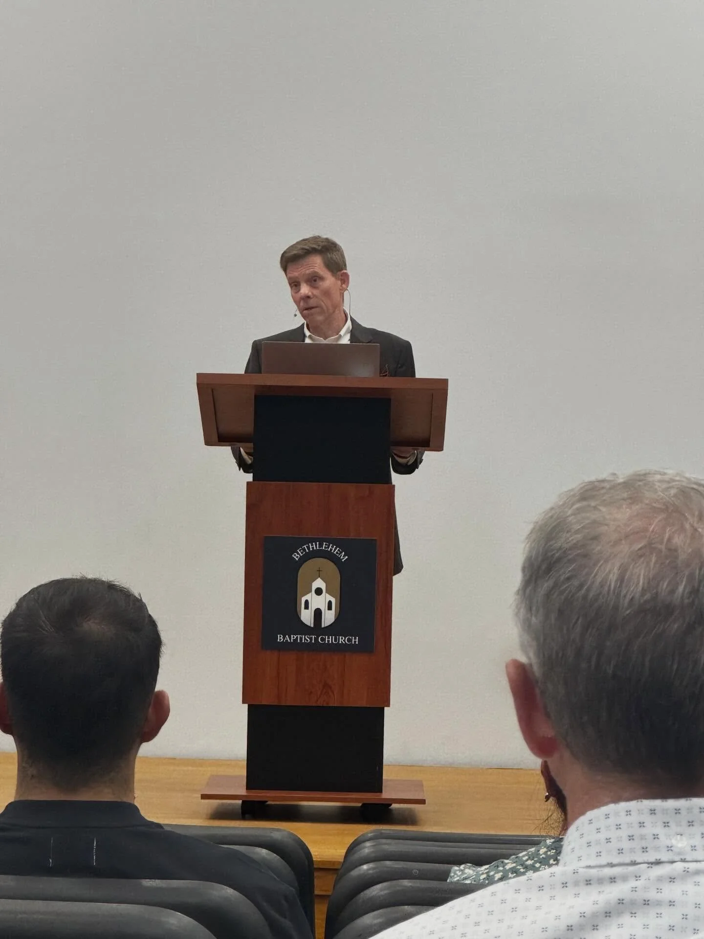 A man in a suit stands at a podium in front of a plain white wall, speaking or giving a presentation at Bethlehem Baptist Church. Several audience members are visible from the back.