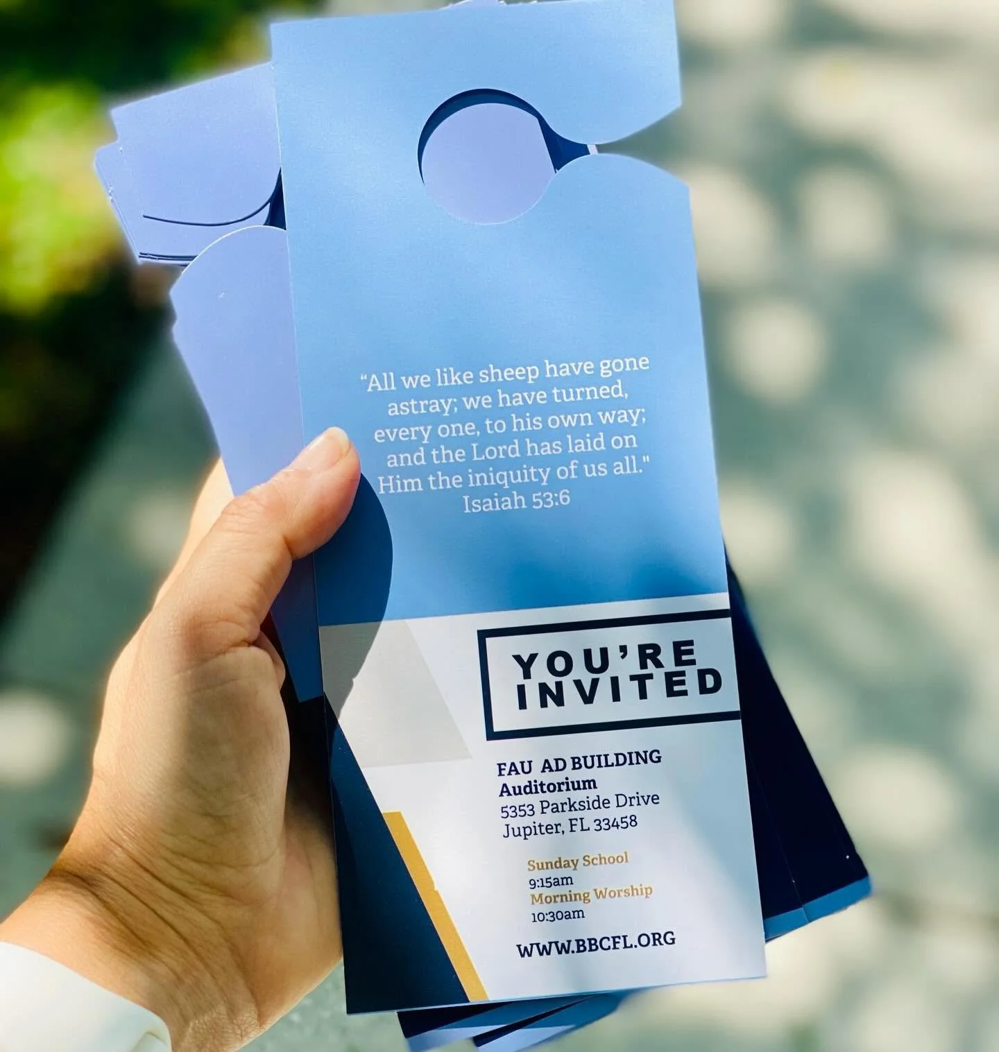 A person holding a church event flyer with a Bible verse at the top, event location, time, and website URL, outdoors.