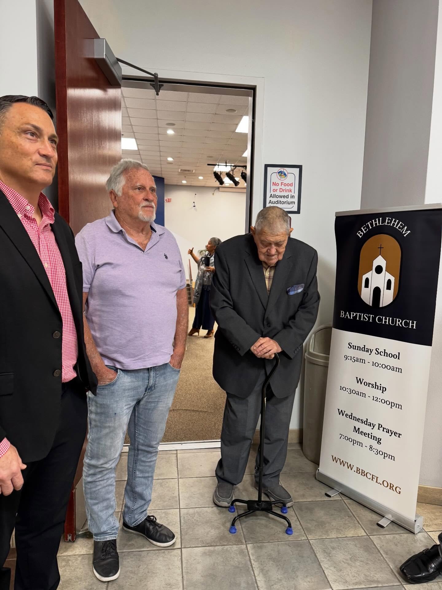 Three men standing near a signboard for Bethlehem Baptist Church, with a fourth person visible in the background. The sign provides the schedule for Sunday School, Worship, and Wednesday Prayer Meetings. An elderly man with a cane is standing with bowed head.