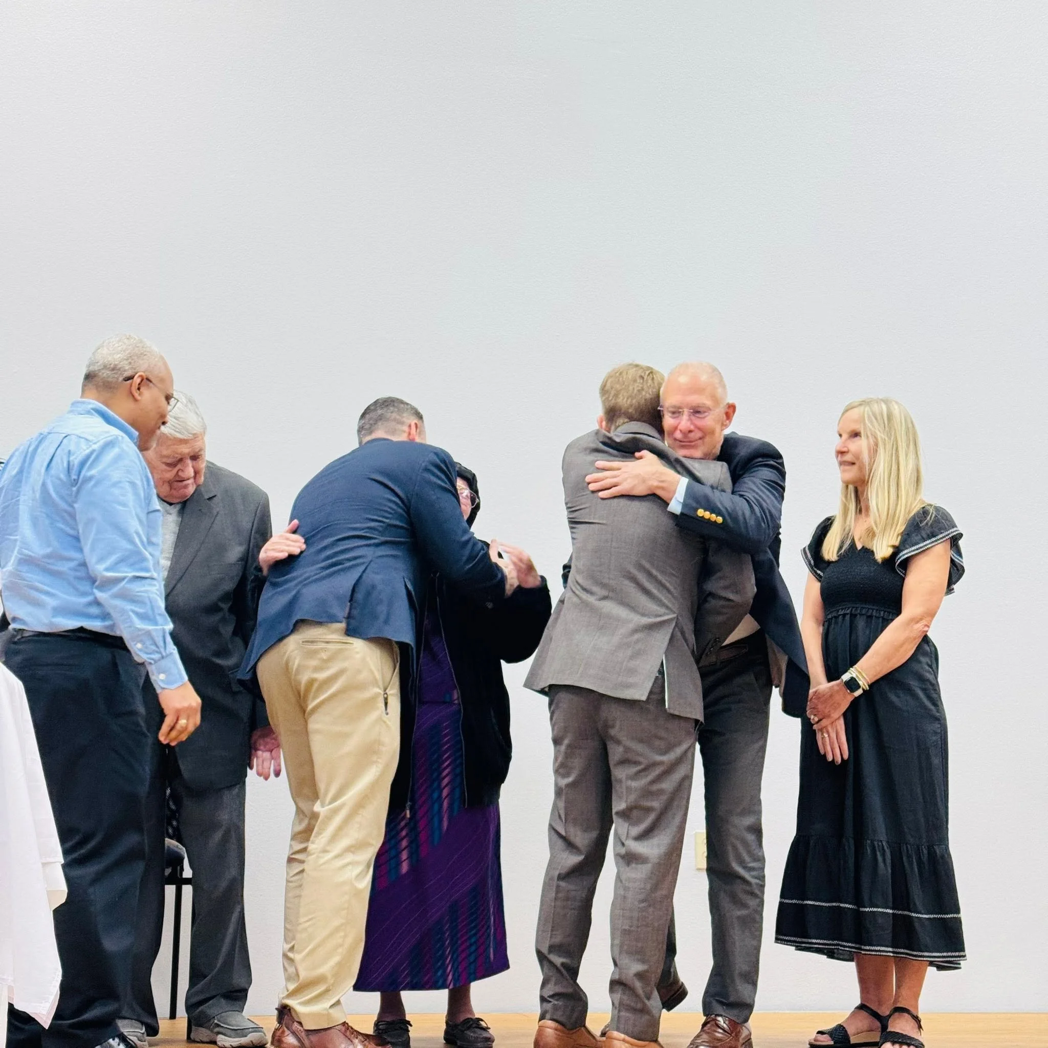 Group of people gathered in a meeting or event, engaging in conversations and embracing each other, in a room with white walls.