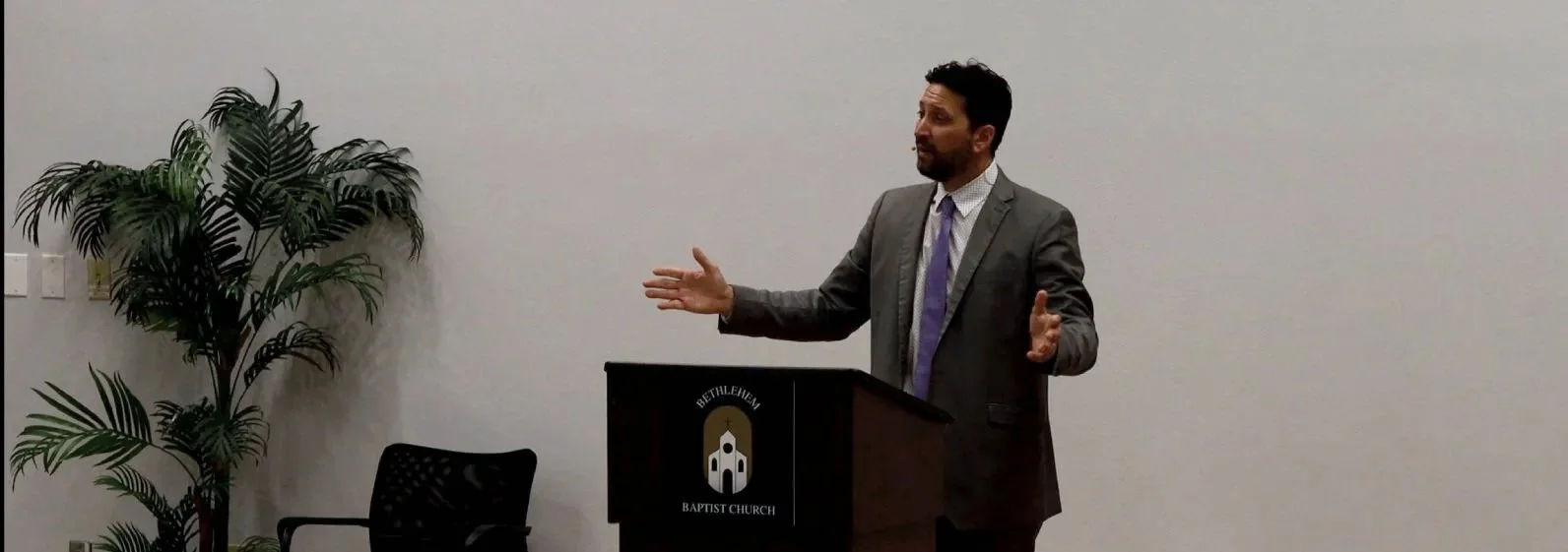 A man in a gray suit and purple tie speaking at a podium at Bethlehem Baptist Church, with a potted plant on the left side of the image.
