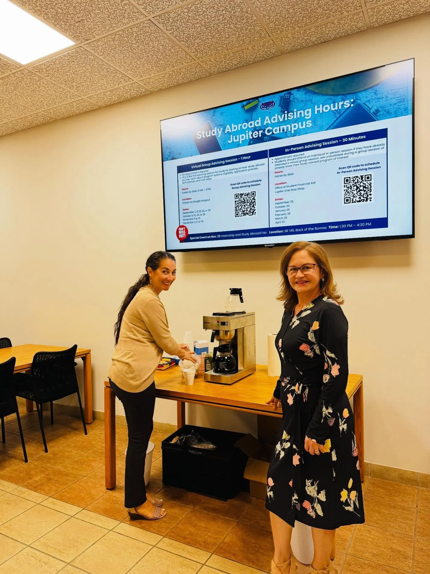 Two women smiling at a coffee station in a room with beige walls and a large screen on the wall displaying study abroad advising hours and details, with chairs and tables around.