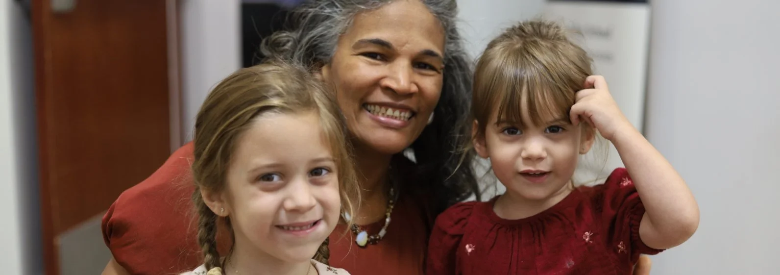 A woman with gray hair smiling with two young girls, one with braids and the other with bangs, in a close-up indoor setting.