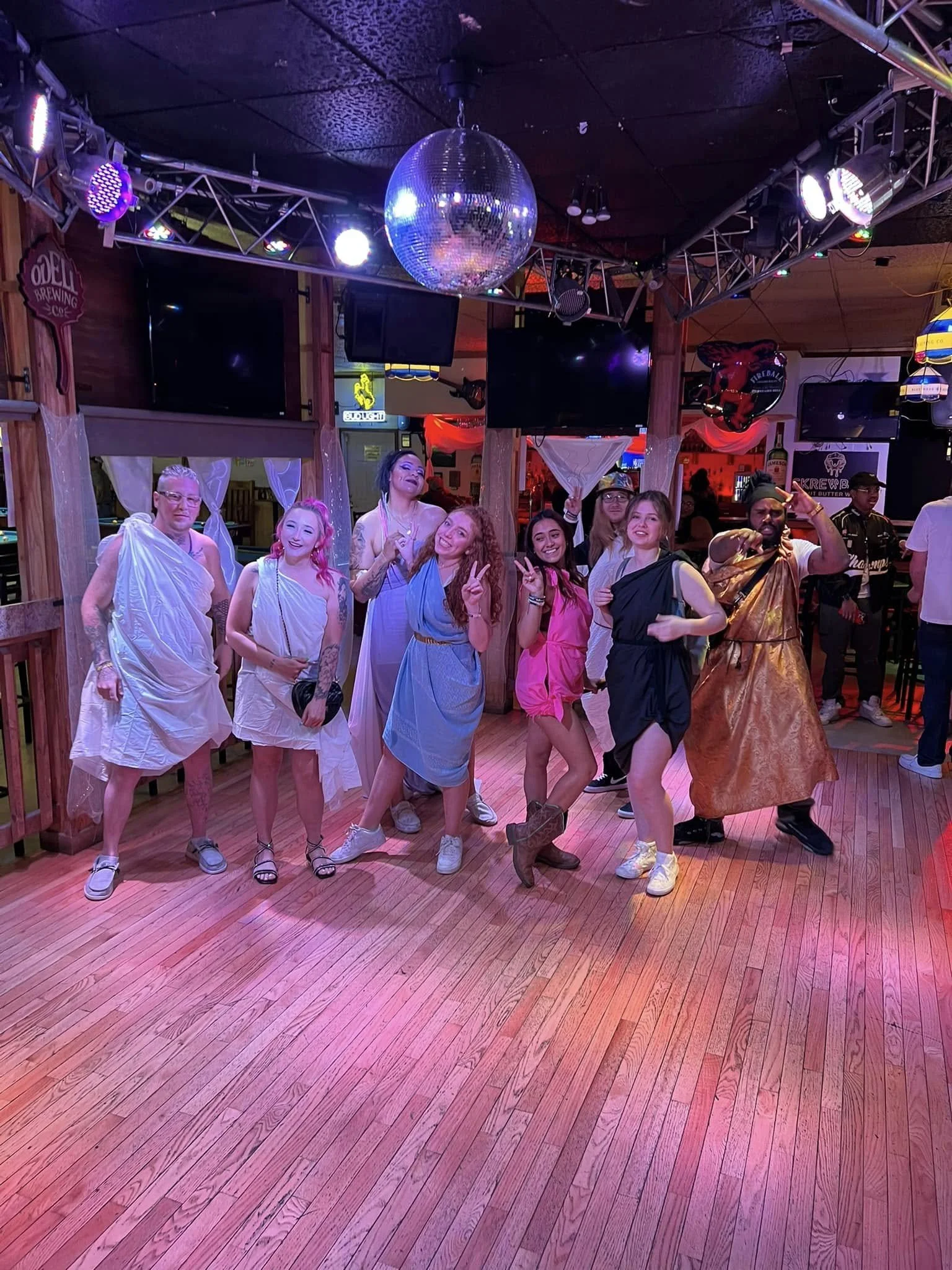 Group of people in colorful costumes posing together inside a bar or club with wooden floors, large TV screens, and a disco ball hanging from the ceiling.
