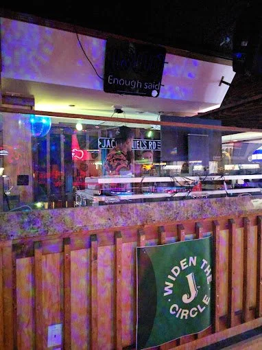 View of an indoor bar or nightclub scene with a disco ball, colorful lighting, and a sign reading 'JACQUES R. S.' in the background. There is a green sign with white text that says 'WIDEN THE CIRCLE' in the foreground.
