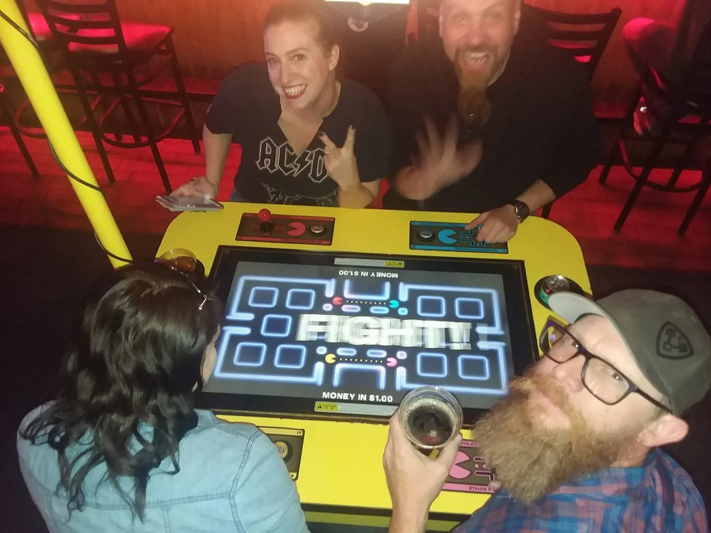 Four people playing a game of digital Pac-Man at a bar or arcade, with two women sitting across from two men, who are celebrating and smiling. One man is holding a drink, and the game screen displays the word 'FIGHT!' in the center.