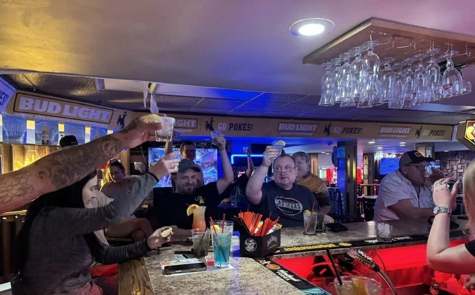 People gathered at a bar raising glasses for a toast, with drinks and bar decor visible.