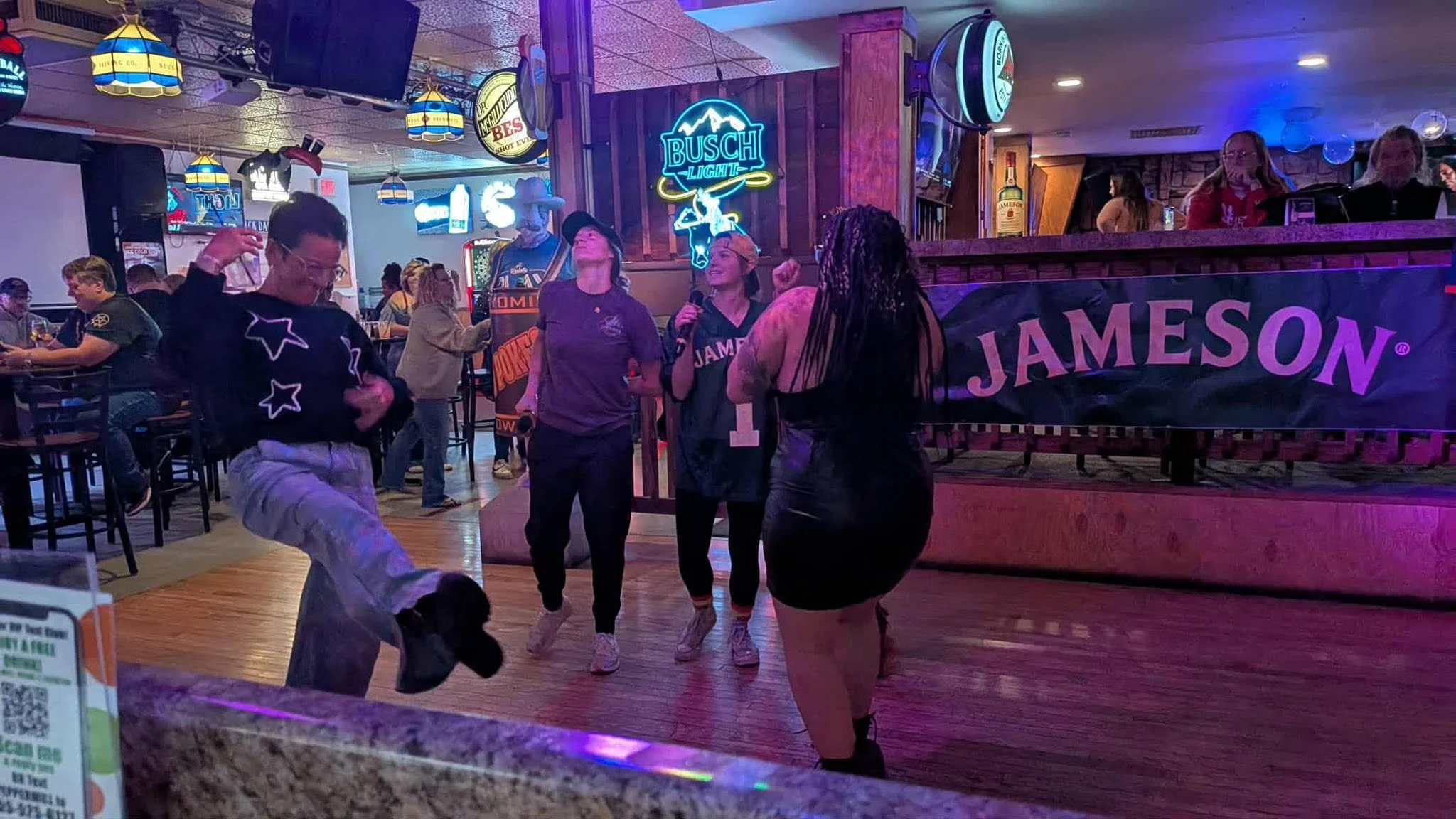 People dancing and socializing in a lively bar or restaurant with neon signs and wooden decor, some wearing masks and casual clothing.