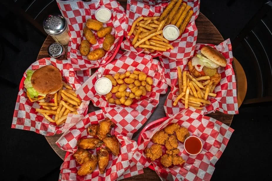 A round wooden table with various fried foods including chicken wings, French fries, onion rings, chicken nuggets, sandwiches, and buffalo wings, all on red and white checkered paper with dipping sauces.