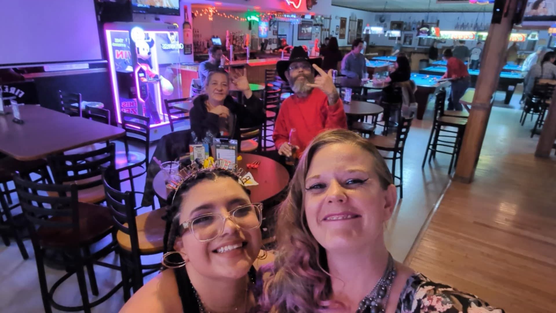 Group selfie at a lively arcade or entertainment center with neon lights, arcade games, and pool tables in the background, featuring smiling people enjoying their time.