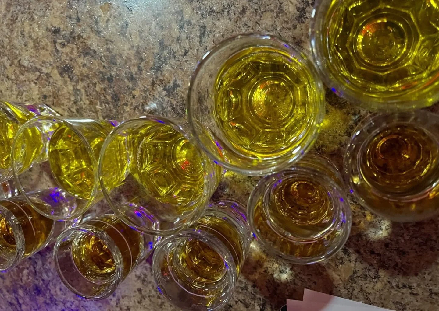 Top-down view of numerous clear glasses filled with yellowish liquid, arranged on a granite or marble countertop.