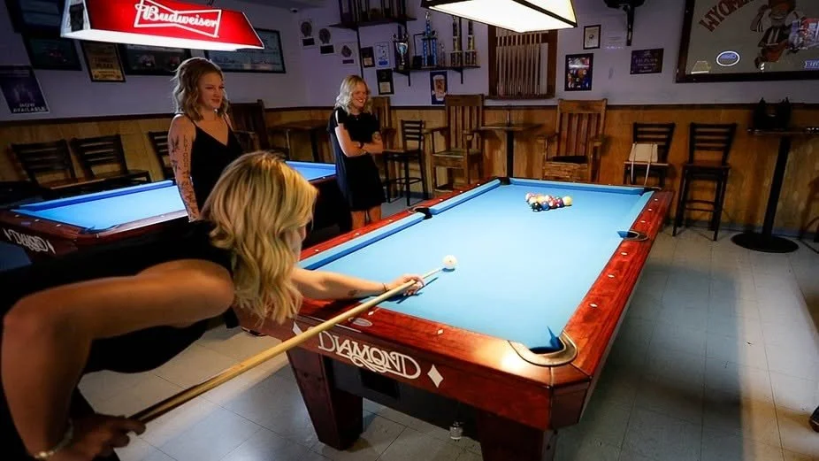 Three women with blonde hair in a bar playing pool, one is lining up a shot, while the other two watch, with bar stools and shelves in the background.