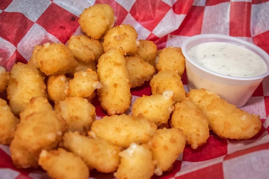 Fried cheese curds served with ranch dipping sauce in a small cup, on red and white checkered paper.