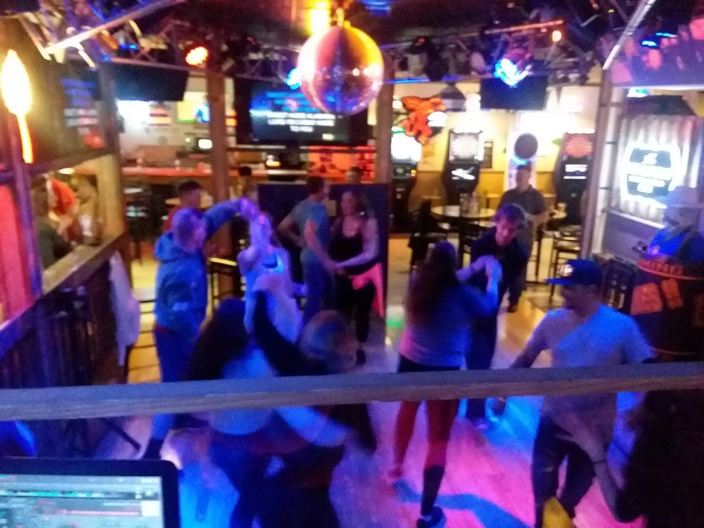 People dancing and socializing in a dimly lit bar or nightclub with colorful neon lights, TV screens, and dartboards on the walls.