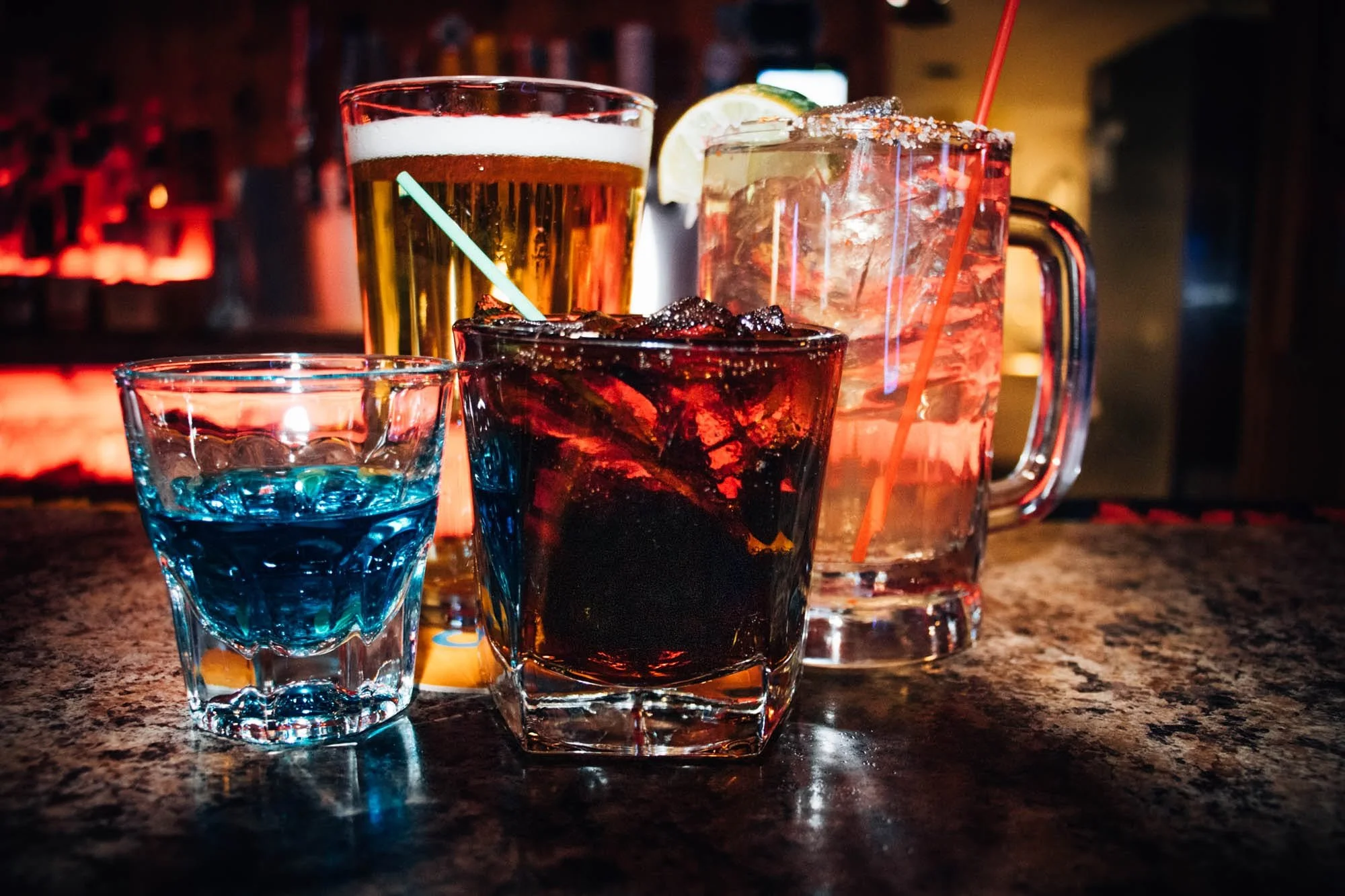Assorted colorful drinks on a bar counter, including a small blue shot glass, a glass of red beverage with ice, a tall glass of beer, and a large glass of mixed drink with lime garnish. The background is dimly lit with bottles and a neon glow.