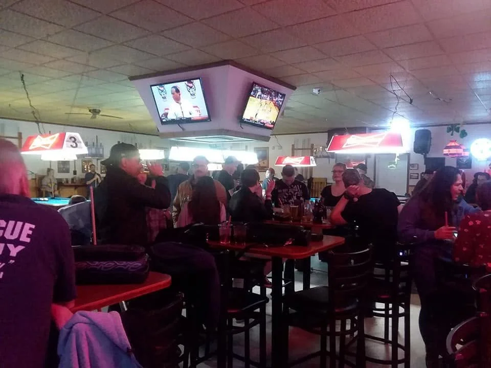 People gathered around tables at a bar or pub, watching sports on hanging TVs, with a pool table in the background and various neon signs on the wall.