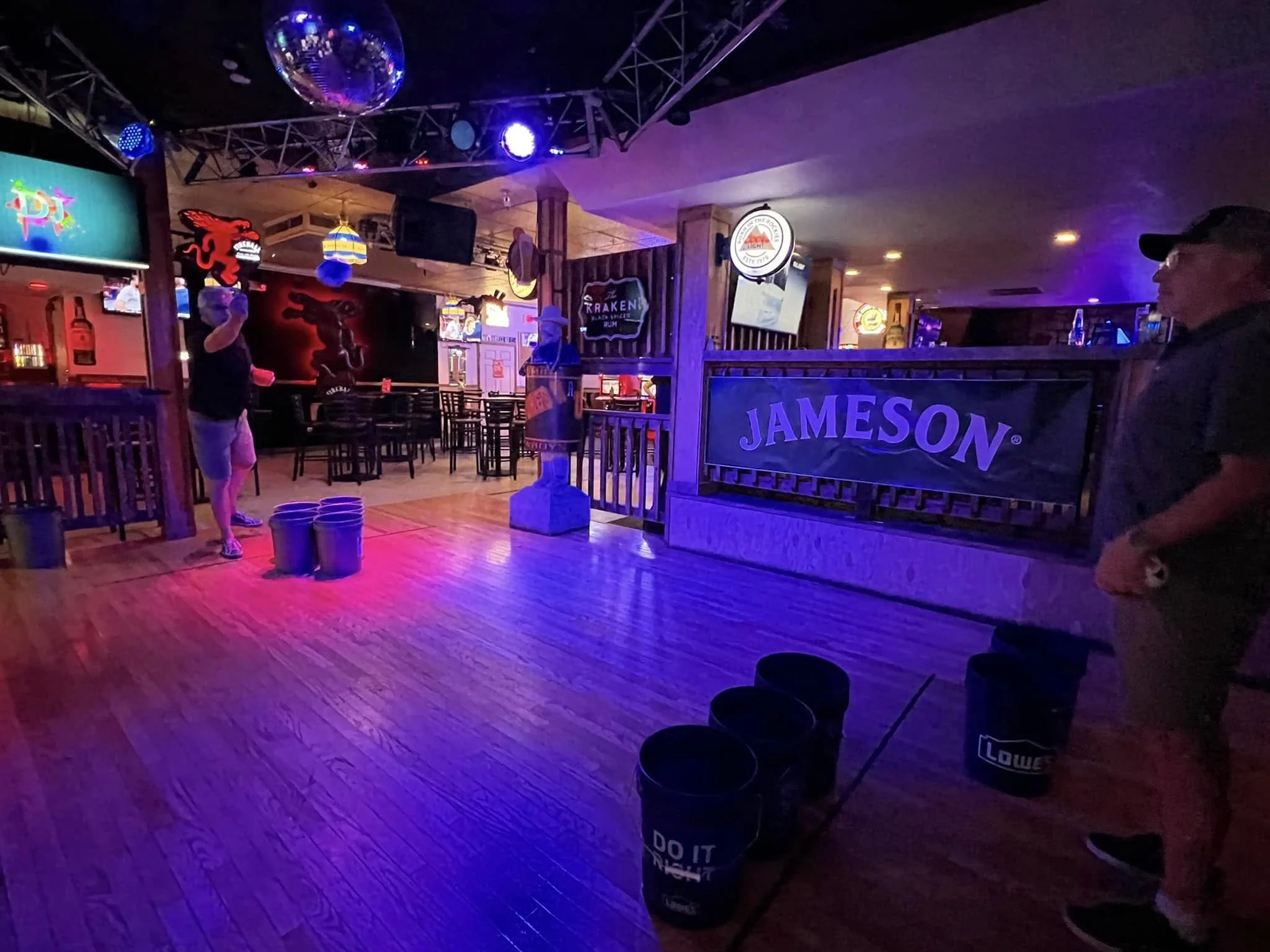 A dimly lit bar with neon signs and a wooden dance floor. There are tables and chairs, and a person is visible holding a drink near the center. Neon lights and signs with a red dragon and drink advertisements enhance the lively atmosphere.