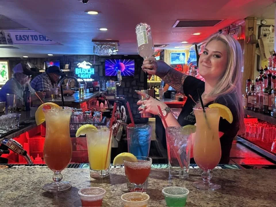 A woman with blonde hair behind a bar counter, holding a clear bottle of liquor, with colorful cocktails and garnishes in front of her, in a lively bar with neon signs and other patrons in the background.