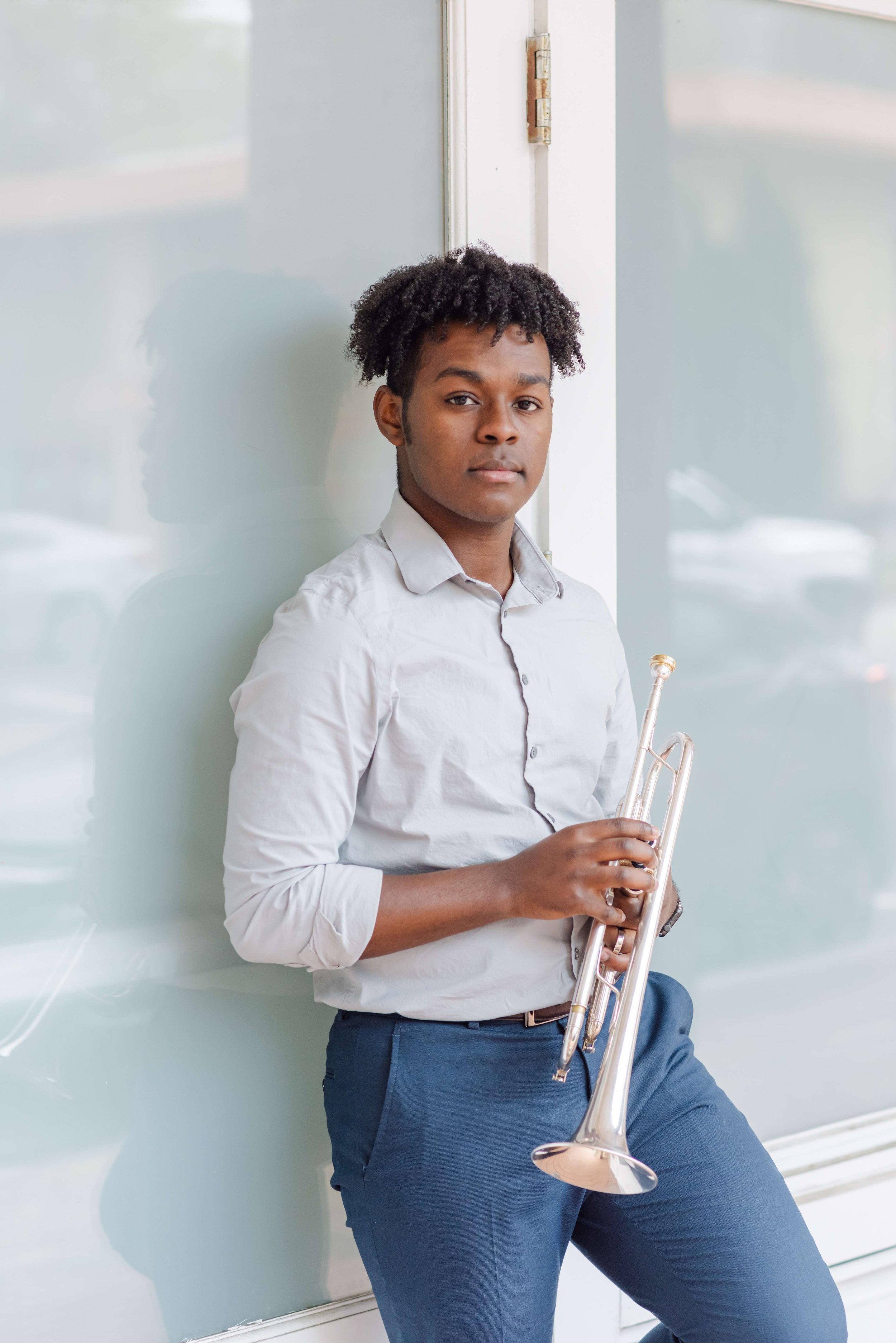 A young man with curly black hair, wearing a white button-up shirt and navy blue pants, holding a trumpet, standing against a light blue wall.