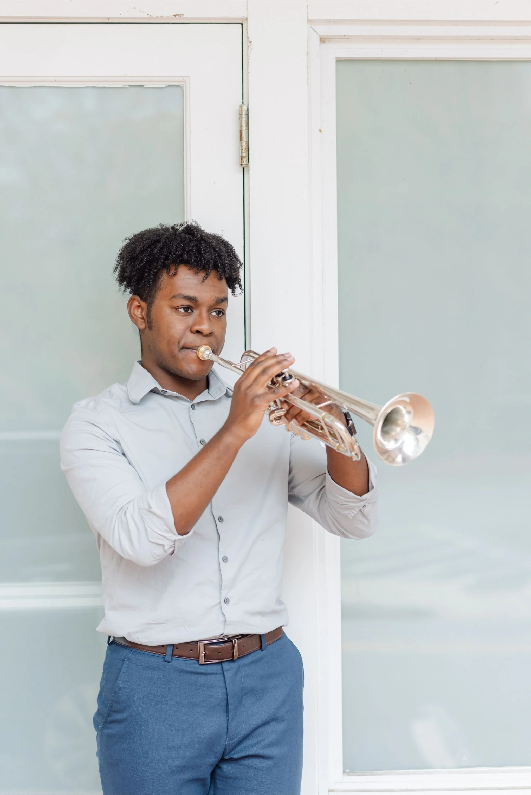 A man with curly black hair wearing a light gray shirt playing a trumpet in front of a white wall and door.