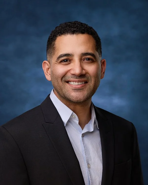 Professional headshot of a smiling man in a black suit and white shirt against a blue background.