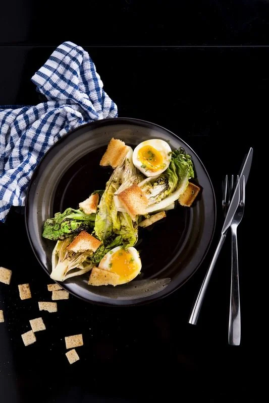A black plate with a salad featuring romaine lettuce, toasted bread cubes, and halved soft-boiled eggs on a black surface with a blue checkered napkin and silver fork and knife nearby.