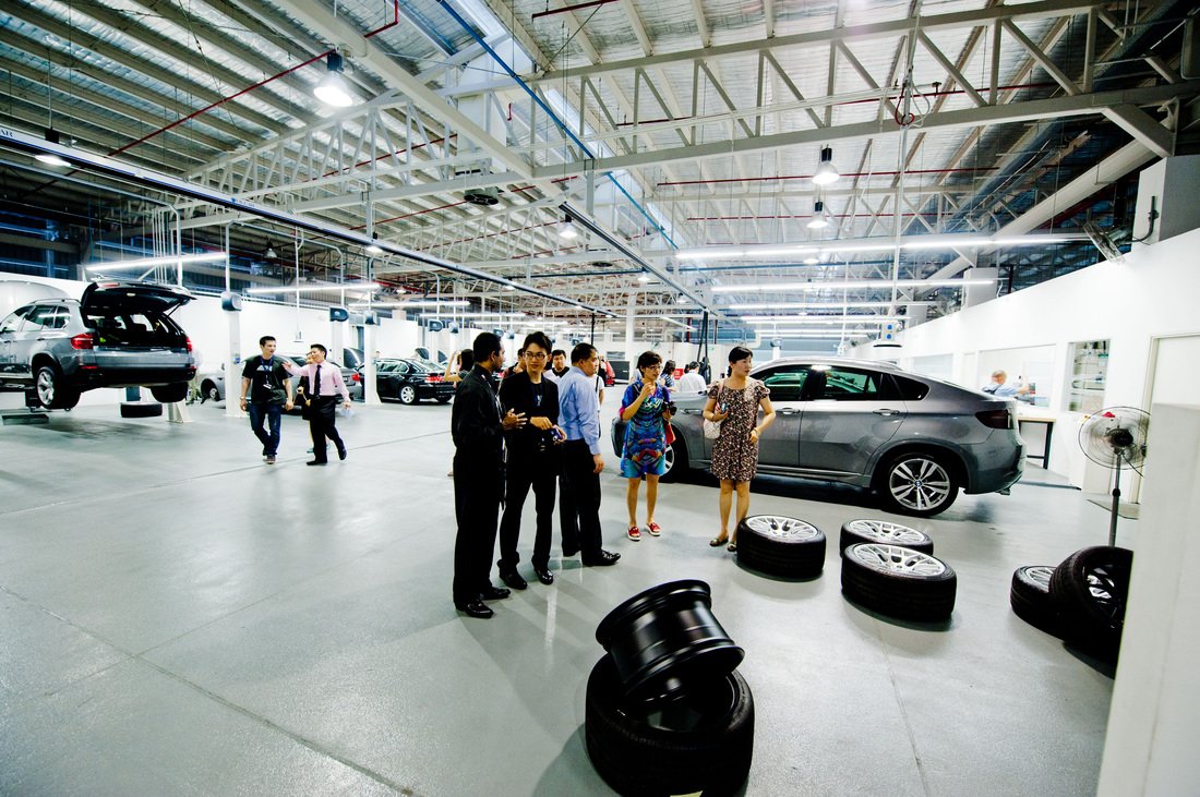 People visiting a car dealership or service center, looking at tires and cars inside a spacious, well-lit industrial building with cars on lifts and floors.