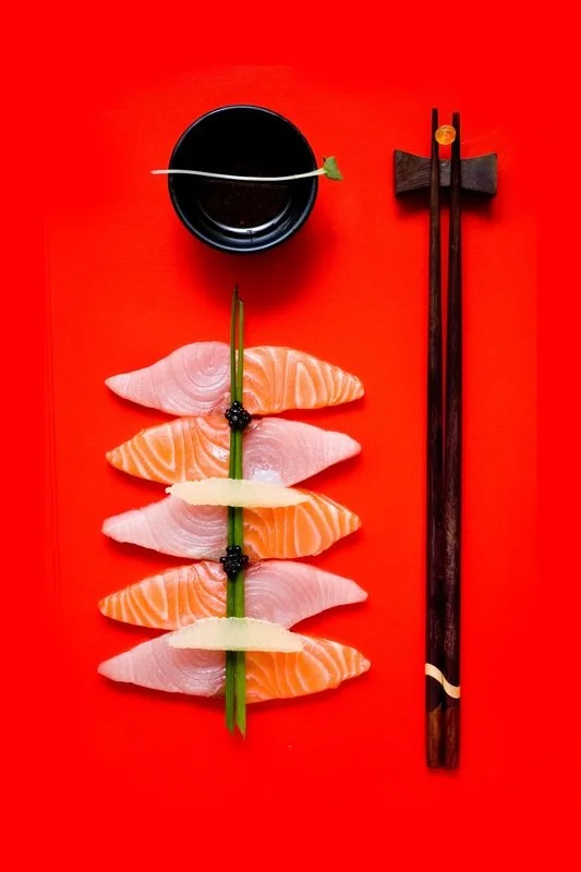 Sliced salmon sashimi arranged on a green scallion on a red background, with a small bowl of soy sauce with a wasabi-topped toothpick and pair of wooden chopsticks.