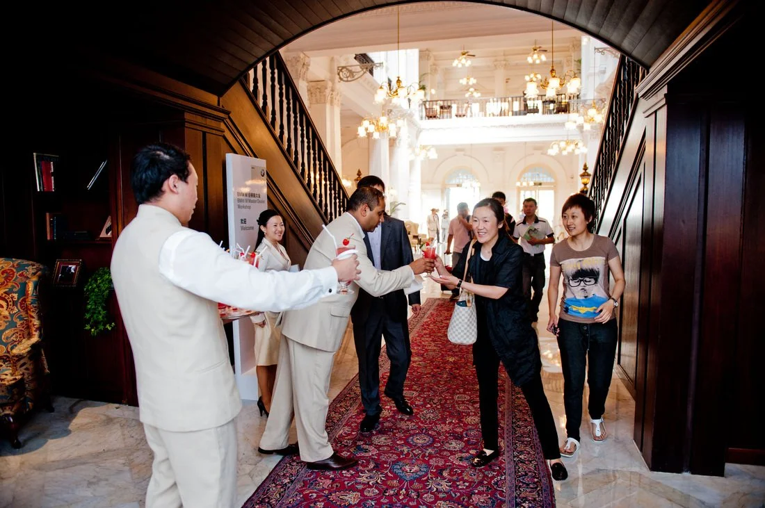 Guests at a hotel check-in counter enjoying drinks and conversation in a luxurious lobby with ornate chandeliers and a staircase.