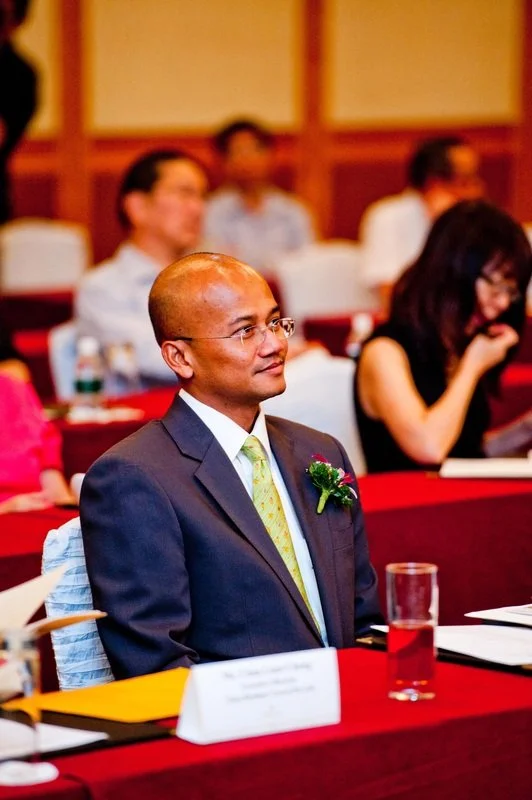 A man in a dark suit and yellow tie sitting at a conference table with a glass of red liquid in front of him, attending a formal event in a decorated room.
