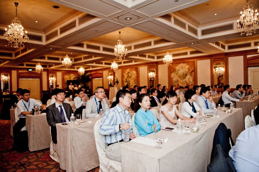 Business conference attendees seated at long tables in a luxurious, chandelier-lit conference room.