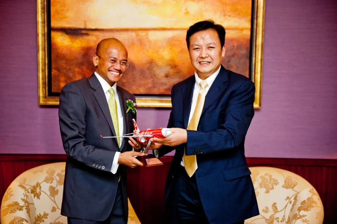 Two men in suits smiling and exchanging a model airplane in a formal setting.