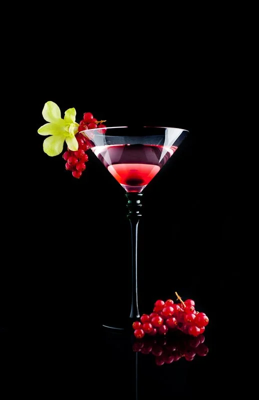A red cocktail in a martini glass garnished with a yellow flower and red berries against a black background.