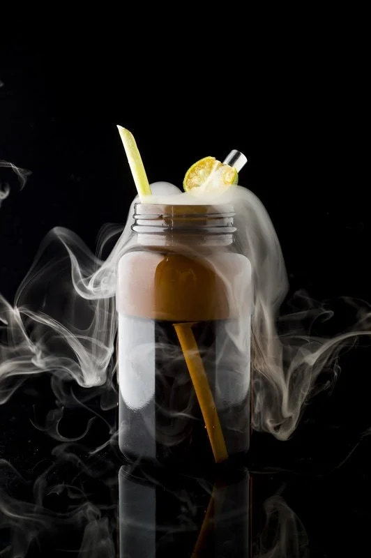 A jar with a cold coffee beverage, lemon slice, and a straw, surrounded by swirling smoke against a black background.