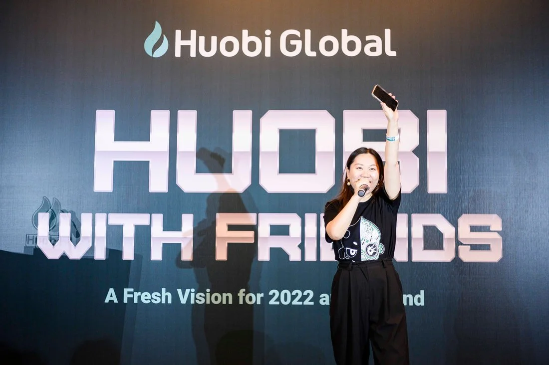 A woman standing on stage holding a microphone and raising her right arm with a phone in hand, in front of a large screen displaying 'Huobi Global' and 'Huobi with Friends' at an event.