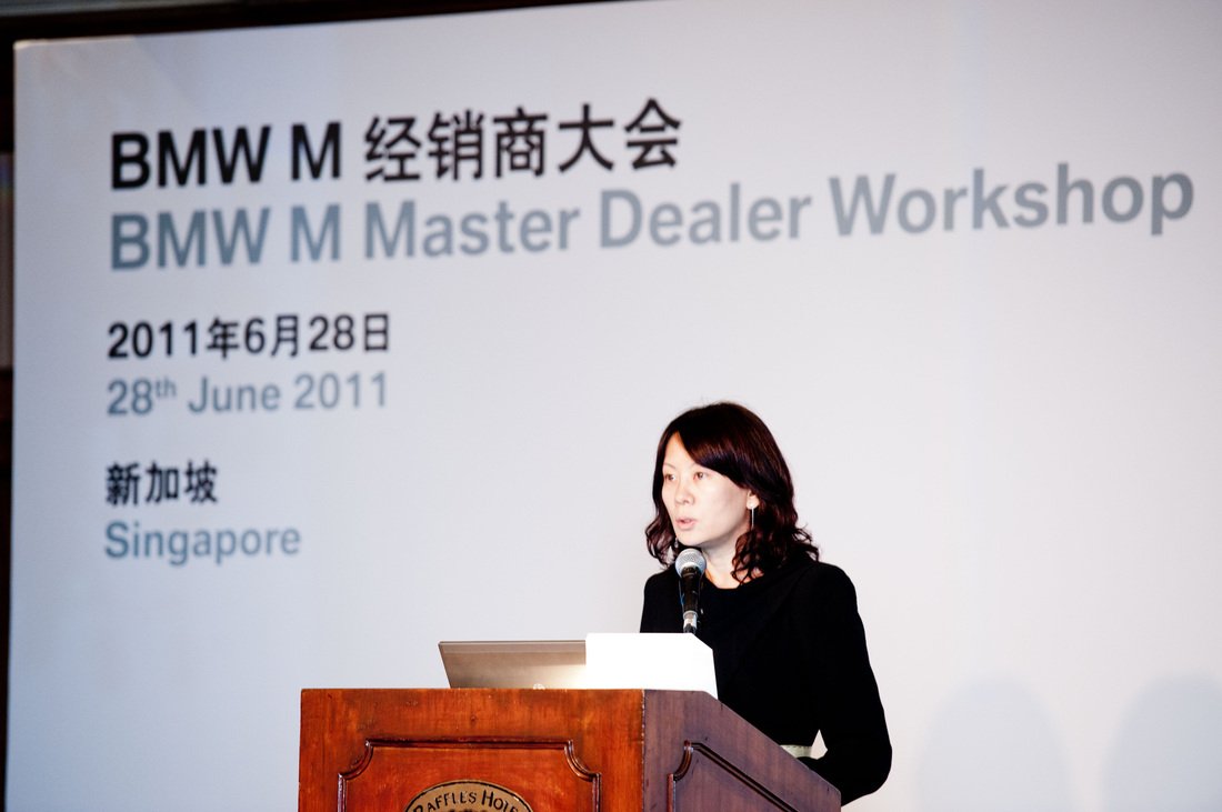 A woman speaking at a conference podium, with a large screen behind her displaying text about a BMW Master Dealer Workshop held in Singapore on June 28, 2011.